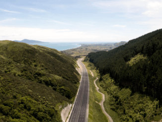Aerial view of Transmission Gully motorway winding through green hills toward the Kāpiti Coast, with the sea visible in the distance.