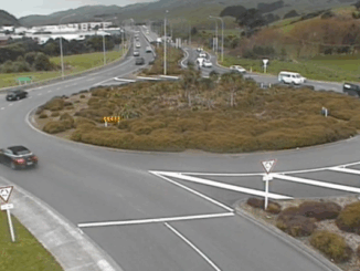 Traffic moves through a large multi-lane roundabout with cars entering and exiting from several directions, surrounded by low shrubs and rolling hills.