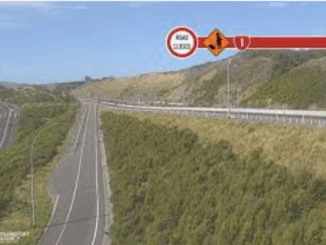 A stretch of Transmission Gully motorway at Waitangirua winds through green hills, with electronic road signs indicating a temporary 50km/h speed limit and lane closure ahead.