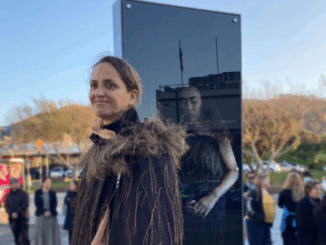 Artist Kohai Grace stands beside a tall glass lightbox artwork during the Te Manawa lightbox unveiling in Porirua in 2022, with people gathered in the background.