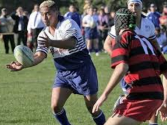 A young rugby player in a blue and white uniform passes the ball during a match, with defenders closing in and spectators watching from the sideline.