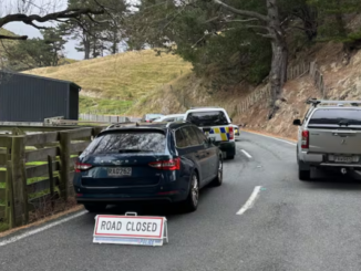 Police vehicles block a narrow rural section of Mākara Rd, with a “Road Closed” sign placed behind a patrol car. The road curves along a hillside with fencing and trees, while officers attend an incident further ahead.