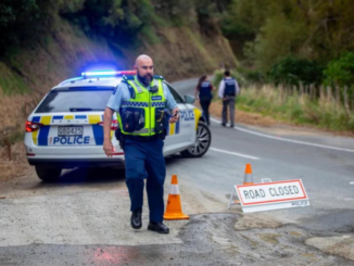 A police officer stands beside a patrol car with flashing lights on a narrow rural road, with cones and a “Road Closed” sign blocking access while other responders work in the background.