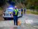 A police officer stands beside a patrol car with flashing lights on a narrow rural road, with cones and a “Road Closed” sign blocking access while other responders work in the background.