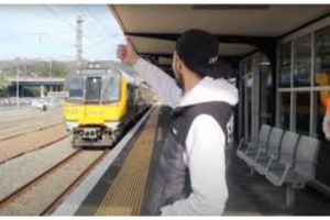 A commuter stands on a train platform raising a hand as a yellow commuter train approaches the station.