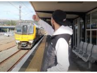 A commuter stands on a train platform raising a hand as a yellow commuter train approaches the station.