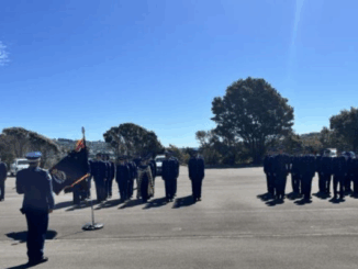 New Zealand Police recruits stand in formation during a graduation parade at the Royal New Zealand Police College in Porirua, with a flag bearer and officer addressing the wing as families watch nearby.