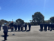 New Zealand Police recruits stand in formation during a graduation parade at the Royal New Zealand Police College in Porirua, with a flag bearer and officer addressing the wing as families watch nearby.