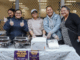 Five women stand behind a food stall at a Pacific night market, smiling and giving thumbs up beside trays of island food.