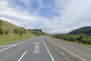 A divided section of State Highway 58 in Porirua, with two lanes in each direction separated by flexible median posts, curving through green hills under a partly cloudy sky.