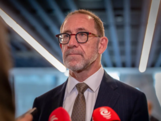 Wellington Mayor Andrew Little speaking to reporters indoors, wearing a suit and glasses, with microphones visible in the foreground.