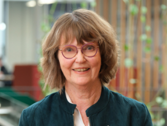 Adrienne Staples is pictured indoors, wearing glasses and a dark jacket, with a softly blurred background. She is smiling in a professional portrait taken during her time in public office.