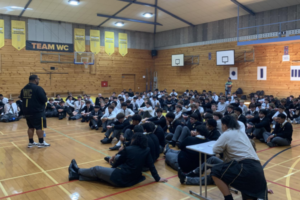A large group of students in school uniform sit on a gymnasium floor, listening to a speaker standing at the front.