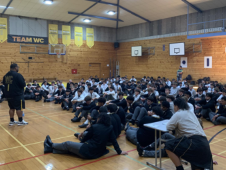 A large group of students in school uniform sit on a gymnasium floor, listening to a speaker standing at the front.