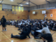 A large group of students in school uniform sit on a gymnasium floor, listening to a speaker standing at the front.