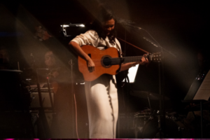 Bic Runga looks down at her acoustic guitar while playing on stage, softly lit, with orchestral musicians and instruments visible in the background.