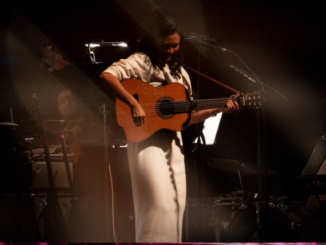 Bic Runga looks down at her acoustic guitar while playing on stage, softly lit, with orchestral musicians and instruments visible in the background.
