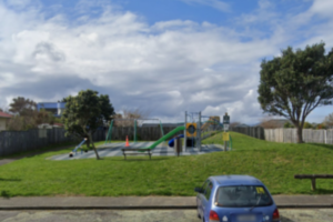 A small neighbourhood playground with a slide, swings and climbing frame sits on a grassy hill, with a parked car visible in the foreground.