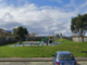 A small neighbourhood playground with a slide, swings and climbing frame sits on a grassy hill, with a parked car visible in the foreground.