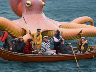 A group of seven people in a small wooden rowboat float on open water beside a giant, brightly coloured inflatable octopus with patterned tentacles and large yellow eyes.