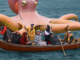 A group of seven people in a small wooden rowboat float on open water beside a giant, brightly coloured inflatable octopus with patterned tentacles and large yellow eyes.