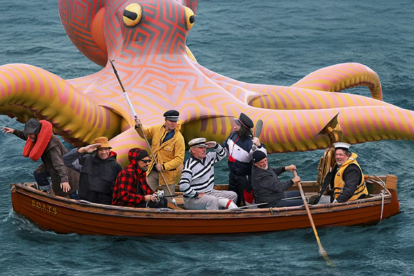 A group of seven people in a small wooden rowboat float on open water beside a giant, brightly coloured inflatable octopus with patterned tentacles and large yellow eyes.