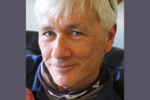 Close-up portrait of an older man with short grey hair and light-coloured eyes, smiling gently at the camera. He is wearing a dark top and a patterned neck scarf, seated indoors against a neutral background.