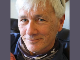 Close-up portrait of an older man with short grey hair and light-coloured eyes, smiling gently at the camera. He is wearing a dark top and a patterned neck scarf, seated indoors against a neutral background.