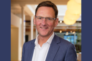 A middle-aged man wearing glasses and a blue blazer smiles at the camera in a well-lit indoor office setting.