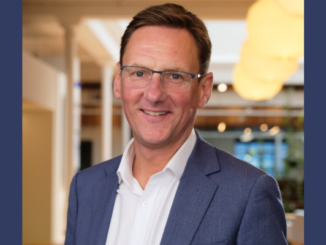 A middle-aged man wearing glasses and a blue blazer smiles at the camera in a well-lit indoor office setting.