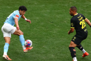 A Wellington Phoenix player in a light blue kit controls the ball mid-air while a Melbourne City opponent in a black kit closes in to defend during an A-League Men match.