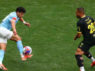 A Wellington Phoenix player in a light blue kit controls the ball mid-air while a Melbourne City opponent in a black kit closes in to defend during an A-League Men match.