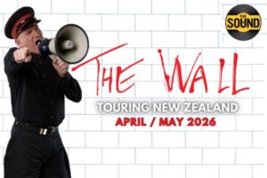 A man dressed in black with a red-trimmed cap shouts into a megaphone against a white brick wall, referencing Pink Floyd’s The Wall. Red graffiti-style text reads “The Wall,” with additional text promoting a New Zealand tour in April and May 2026.