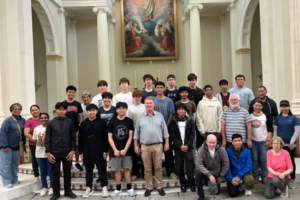 A group of students and adults stand inside a church, posing for a photo in front of an altar with tall white columns and a religious painting above.