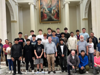 A group of students and adults stand inside a church, posing for a photo in front of an altar with tall white columns and a religious painting above.