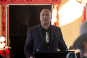 Helmut Modlik stands at a podium speaking to an audience at an indoor event.