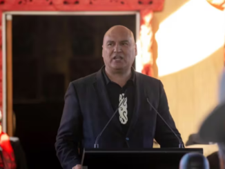 Helmut Modlik stands at a podium speaking to an audience at an indoor event.