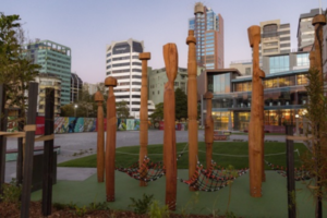 Tall carved wooden tumu sculptures stand in Te Ngākau Civic Square, with modern city buildings rising in the background.