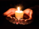 Hands gently cupping a lit candle in a dark setting, symbolising remembrance and mourning.