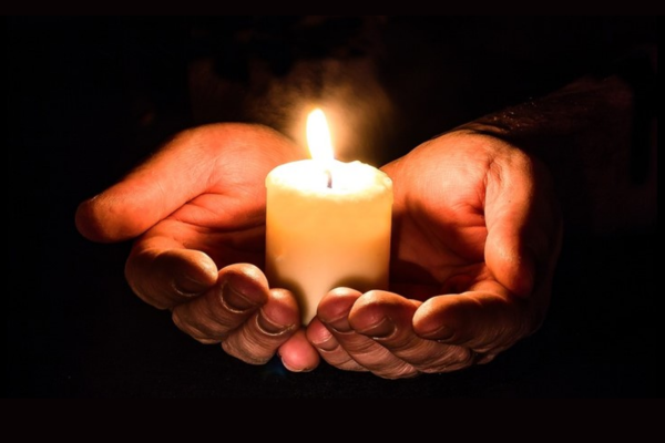 Hands gently cupping a lit candle in a dark setting, symbolising remembrance and mourning.