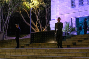 Uniformed service personnel stand guard on the steps of a war memorial at night, illuminated by soft purple and white lighting, with trees and stone walls in the background.