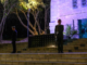 Uniformed service personnel stand guard on the steps of a war memorial at night, illuminated by soft purple and white lighting, with trees and stone walls in the background.