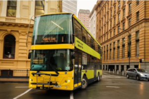 A yellow double-decker Metlink bus drives through a city street lined with historic buildings on a wet day.