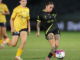 Wellington Phoenix player in black kit strikes the ball during a women’s A-League match as a Brisbane Roar opponent in yellow tracks back.