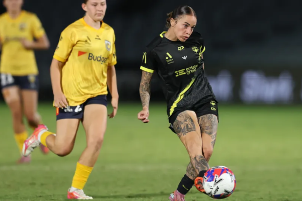 Wellington Phoenix player in black kit strikes the ball during a women’s A-League match as a Brisbane Roar opponent in yellow tracks back.