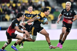 A Hurricanes player in a yellow and black jersey charges forward with the ball at Hnry Stadium in Wellington, breaking through two Crusaders defenders in black and red kits during a Super Rugby Pacific match, while teammates and opponents close in behind.