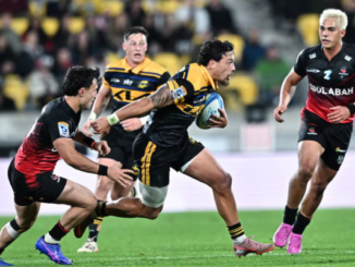 A Hurricanes player in a yellow and black jersey charges forward with the ball at Hnry Stadium in Wellington, breaking through two Crusaders defenders in black and red kits during a Super Rugby Pacific match, while teammates and opponents close in behind.
