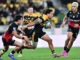 A Hurricanes player in a yellow and black jersey charges forward with the ball at Hnry Stadium in Wellington, breaking through two Crusaders defenders in black and red kits during a Super Rugby Pacific match, while teammates and opponents close in behind.