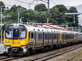 A yellow and silver Metlink train travels along railway tracks beside a Wellington station platform.