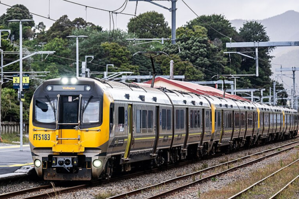 A yellow and silver Metlink train travels along railway tracks beside a Wellington station platform.